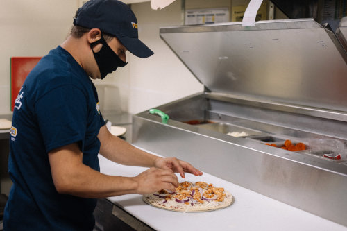 A pizza maker preps a pizza while wearing a mask